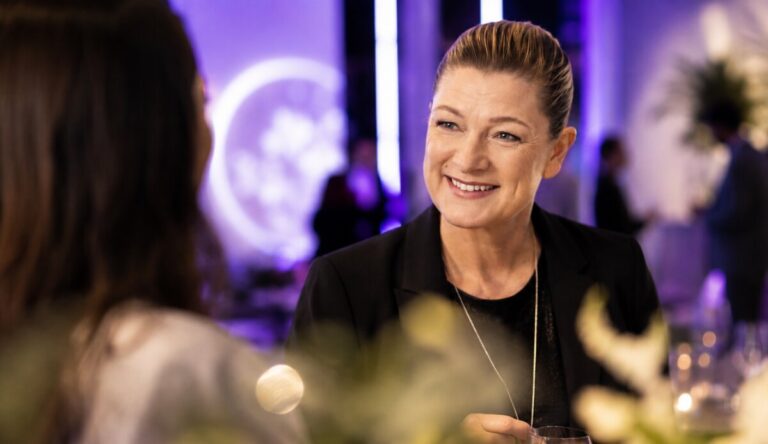 A woman in a black blazer smiles in conversation with a colleague at an evening reception, a glowing moon-like projection on the wall behind her.
