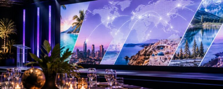 A candlelit banquet table set with glassware and a bronze globe, facing a wide LED wall showing tropical coastlines, a city skyline and a connected world map.
