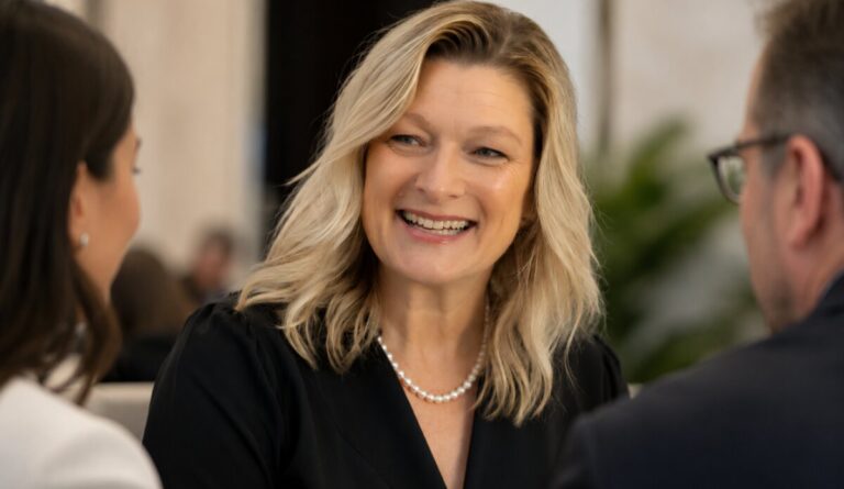 A woman in a black top and pearl necklace smiles warmly in close conversation between two colleagues at an indoor event.