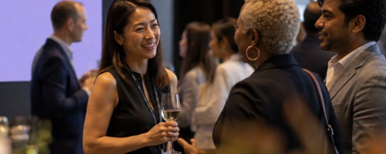 A delegate in a black dress holds a glass of white wine and smiles mid-conversation with two colleagues at a busy evening networking reception.