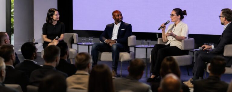 Four panellists in armchairs on a conference stage against a violet screen, one speaking into a handheld microphone while a seated audience watches from the foreground.