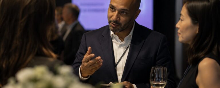 A man in a navy suit and lanyard gestures as he talks with two colleagues over drinks at a conference reception.