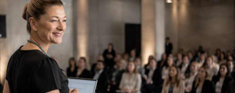 A speaker holding a tablet stands at the front of a full auditorium, looking out over rows of seated guests in a stone-columned hall.