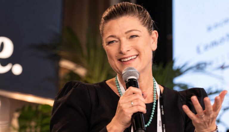 A speaker in a black top and turquoise bead necklace smiles and gestures while answering a question into a handheld microphone on stage.