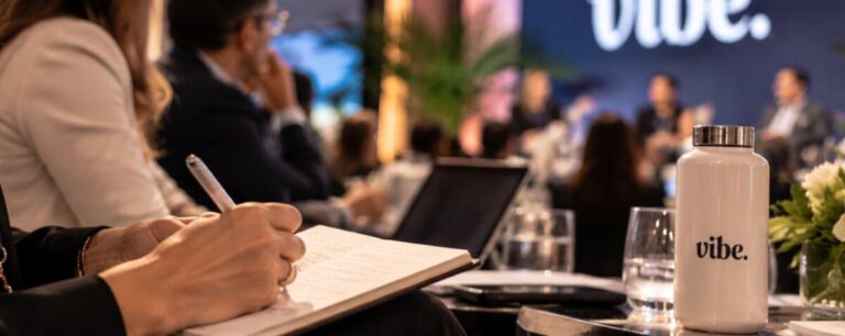 An attendee takes handwritten notes beside a Vibe water bottle during a panel discussion, the speakers blurred on stage beneath a glowing Vibe sign.